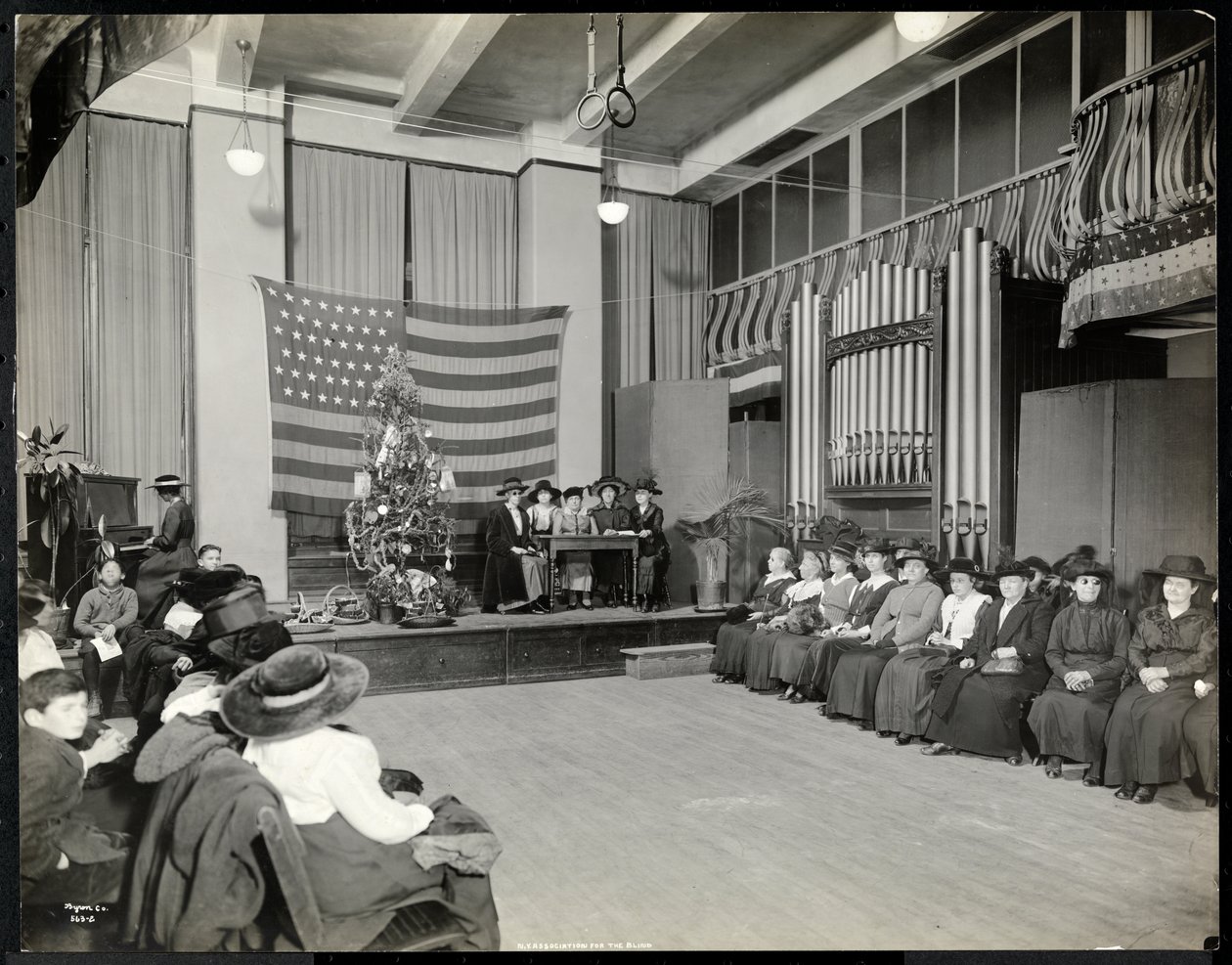 Menschen in einem Auditorium der New York Association for the Blind mit Weihnachtsbaum und möglicherweise einer Aufführung auf der Bühne, 1920 von Byron Company