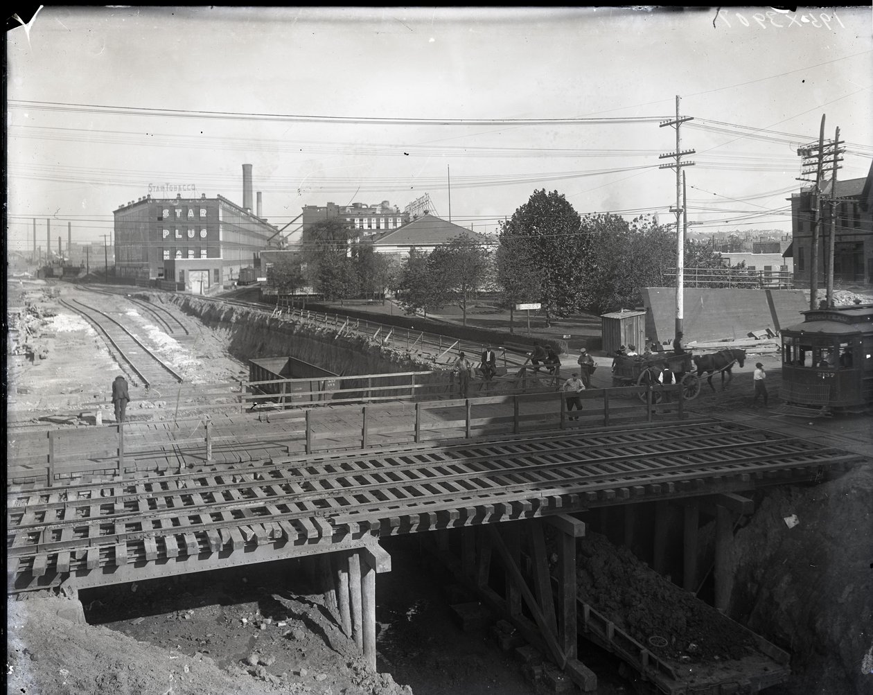 Horizontales Schwarz-Weiß-Foto einer Holzbrücke und Straßenbahnschienen über teilweise gebaute Eisenbahnschienen. Eine Straßenbahn mit der Aufschrift Chouteau 2137 beginnt rechts die Brücke zu überqueren. Im Hintergrund Fabrikgebäude mit der Aufschrift Sta von Charles Clement Holt
