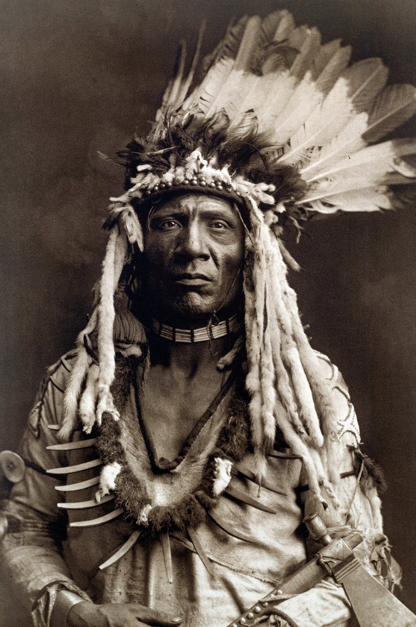 Wiesel Schwanz (Apohsuyis). Porträt des Indianerhäuptlings des Piegan-Stammes, trägt den berühmten Adlerfeder- und Wieselkopfschmuck, eine Wildledertunika, zwei Halsketten, eine aus Knochen und die andere aus Grizzlyklauen, und eine Tomahawk-Pfeife von Edward Sheriff Curtis