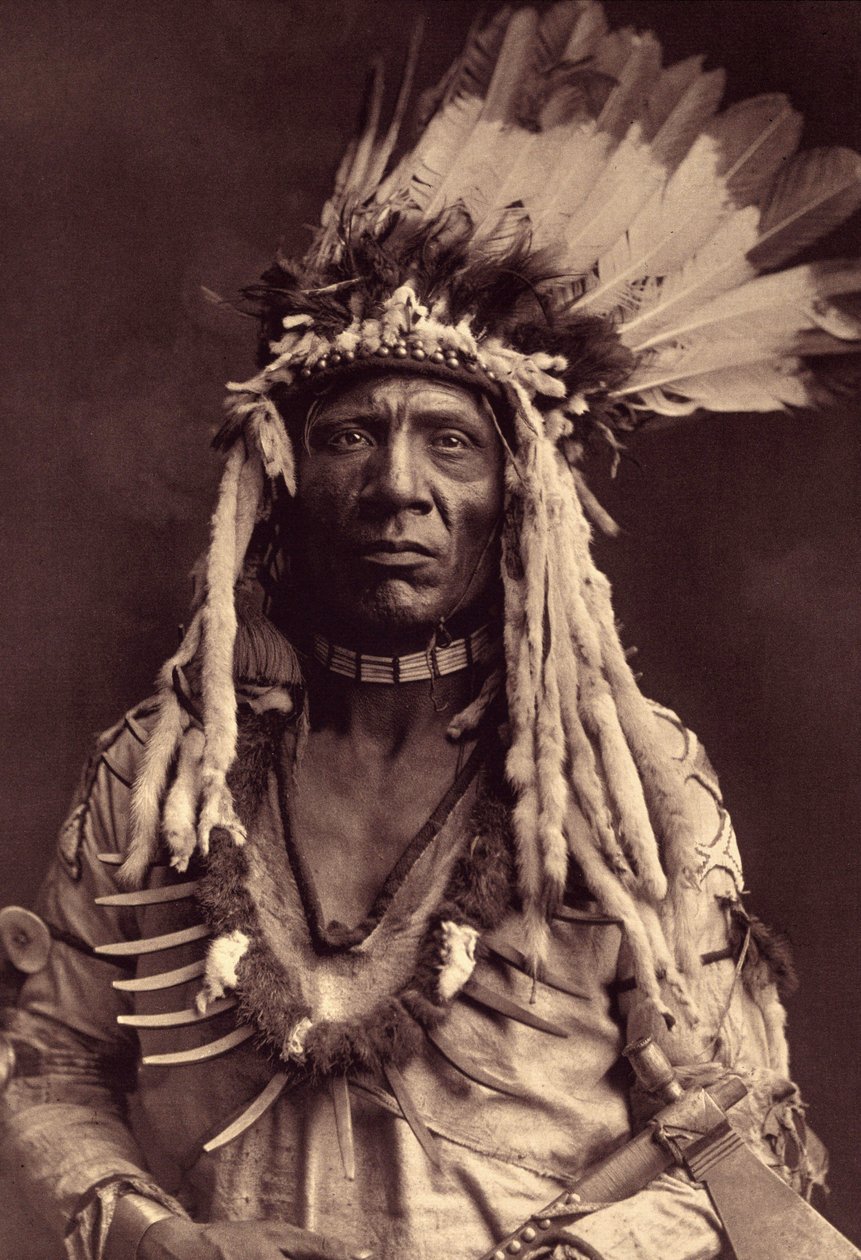 Wiesel-Schwanz (Apohsuyis). Dieser Piegan-Indianer trägt den berühmten Kriegsschmuck aus Adlerfedern und Wieselfell, eine Wildleder-Tunika, zwei Halsketten, eine aus Knochen und die andere aus Grizzly-Klauen, und eine Tomahawk-Pfeife. Foto aus Band 6 der E von Edward Sheriff Curtis