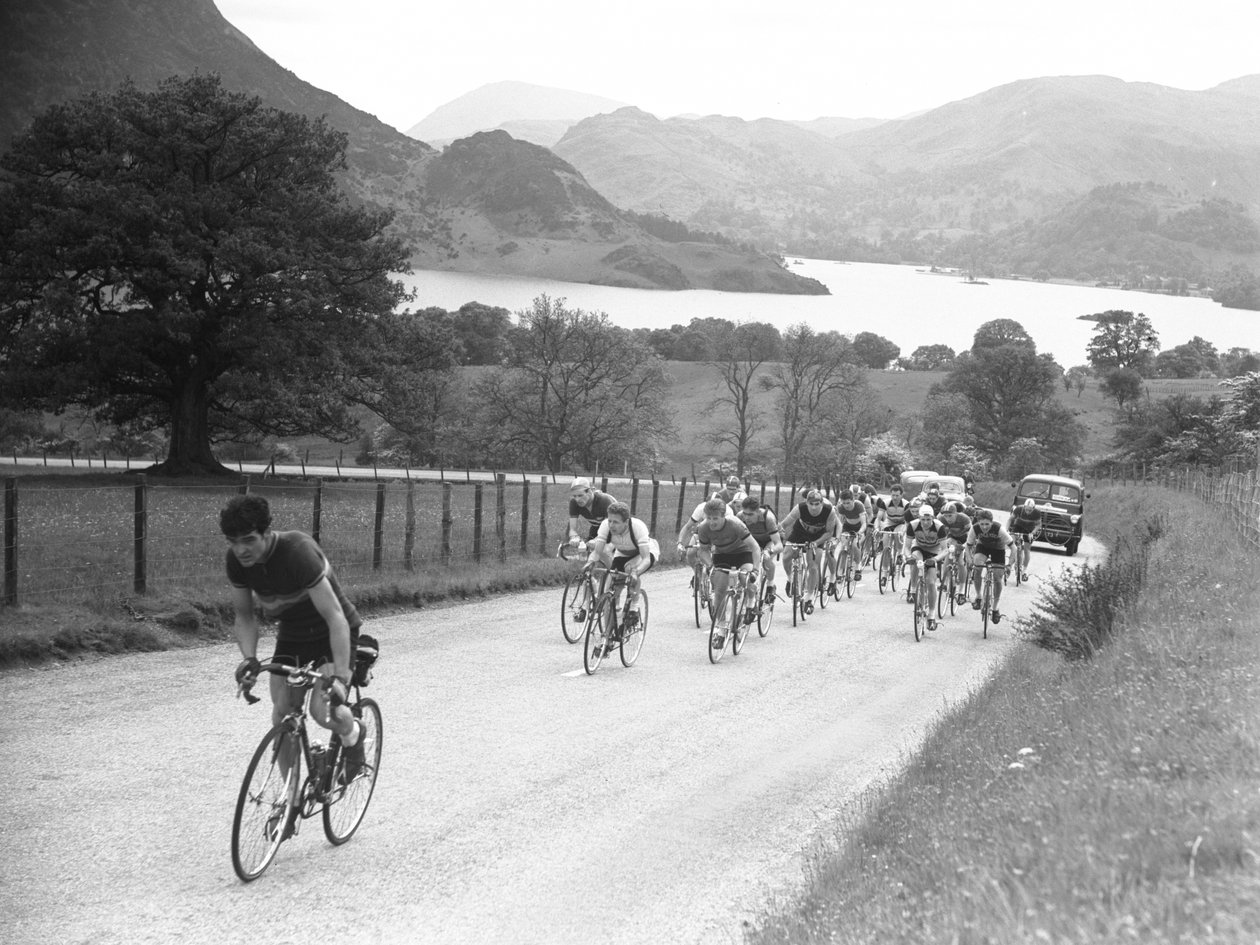 Gruppe von Radfahrern fährt eine Straße hinauf, gefolgt von Autos, See und Hügel im Hintergrund von Joseph Hardman
