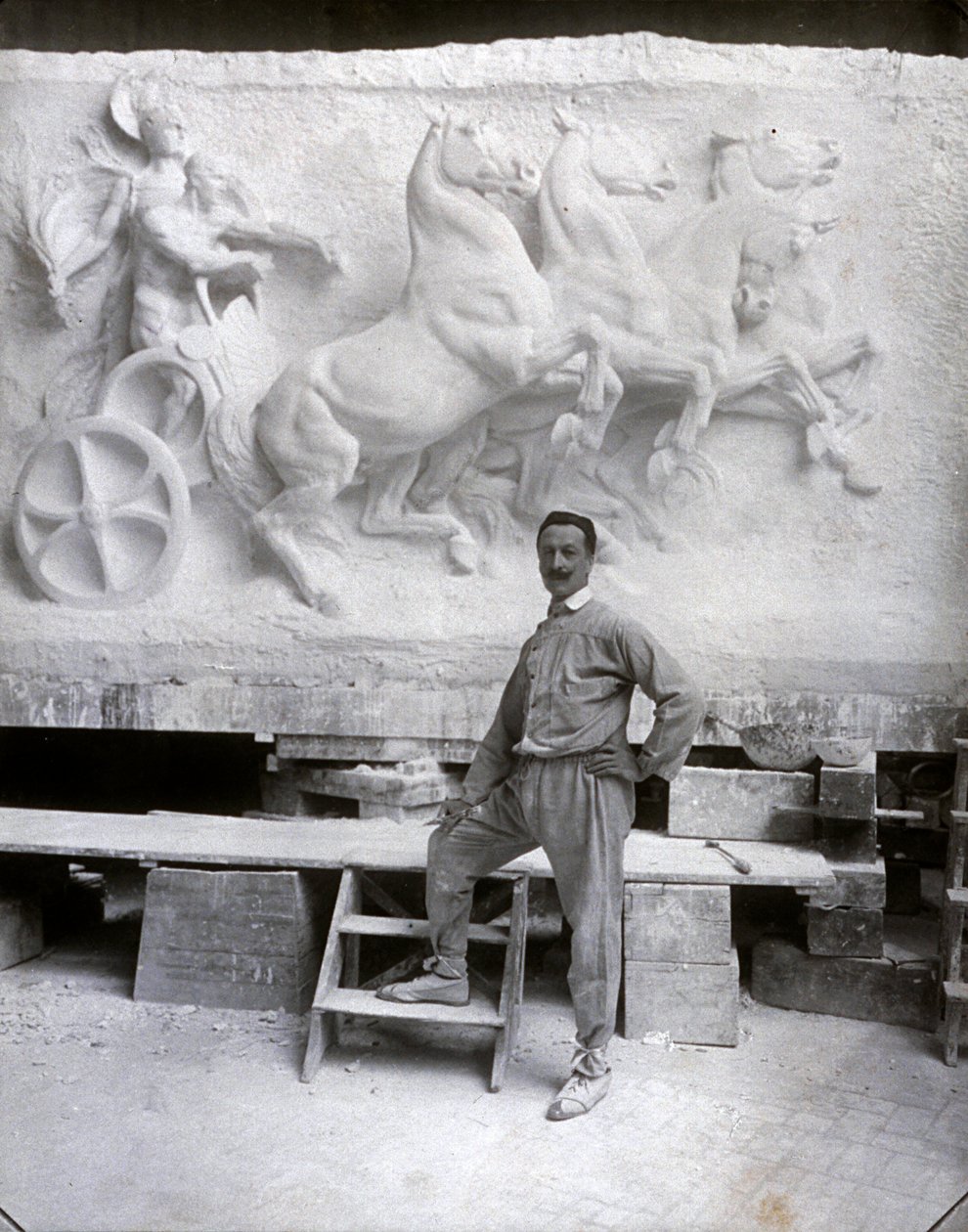 Ganzkörperporträt des berühmten Bildhauers Pietro Canonica. Er posiert in seinem Atelier vor einem Marmorhochrelief, an dem er arbeitet von Mario Nunes Vais