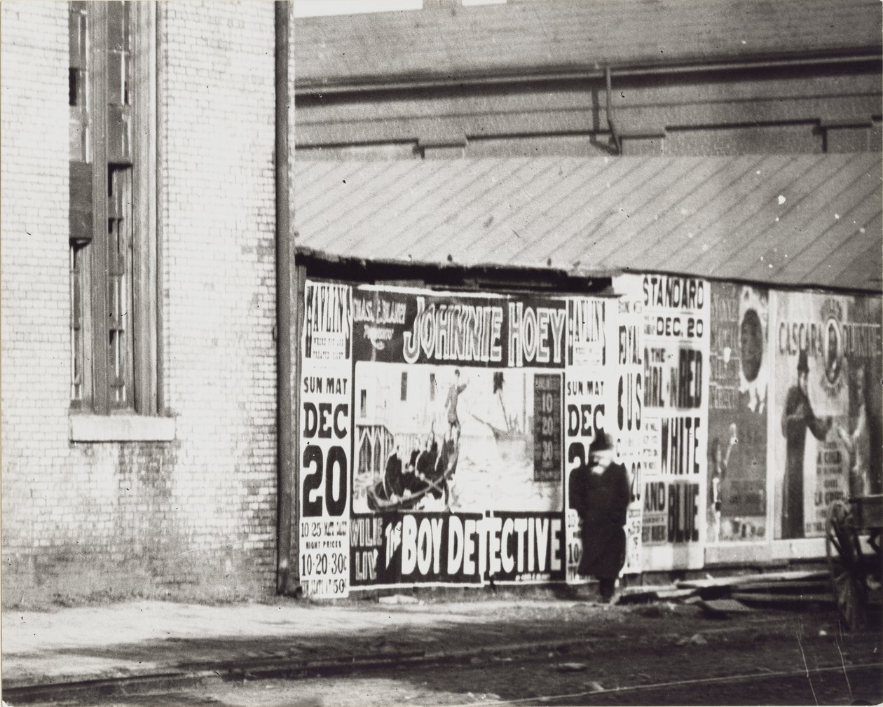 Ein Mann geht an einer Wand mit Plakatwerbung vorbei, darunter eine für Johnnie Hoey, den Jungen Detektiv, der im Havlin