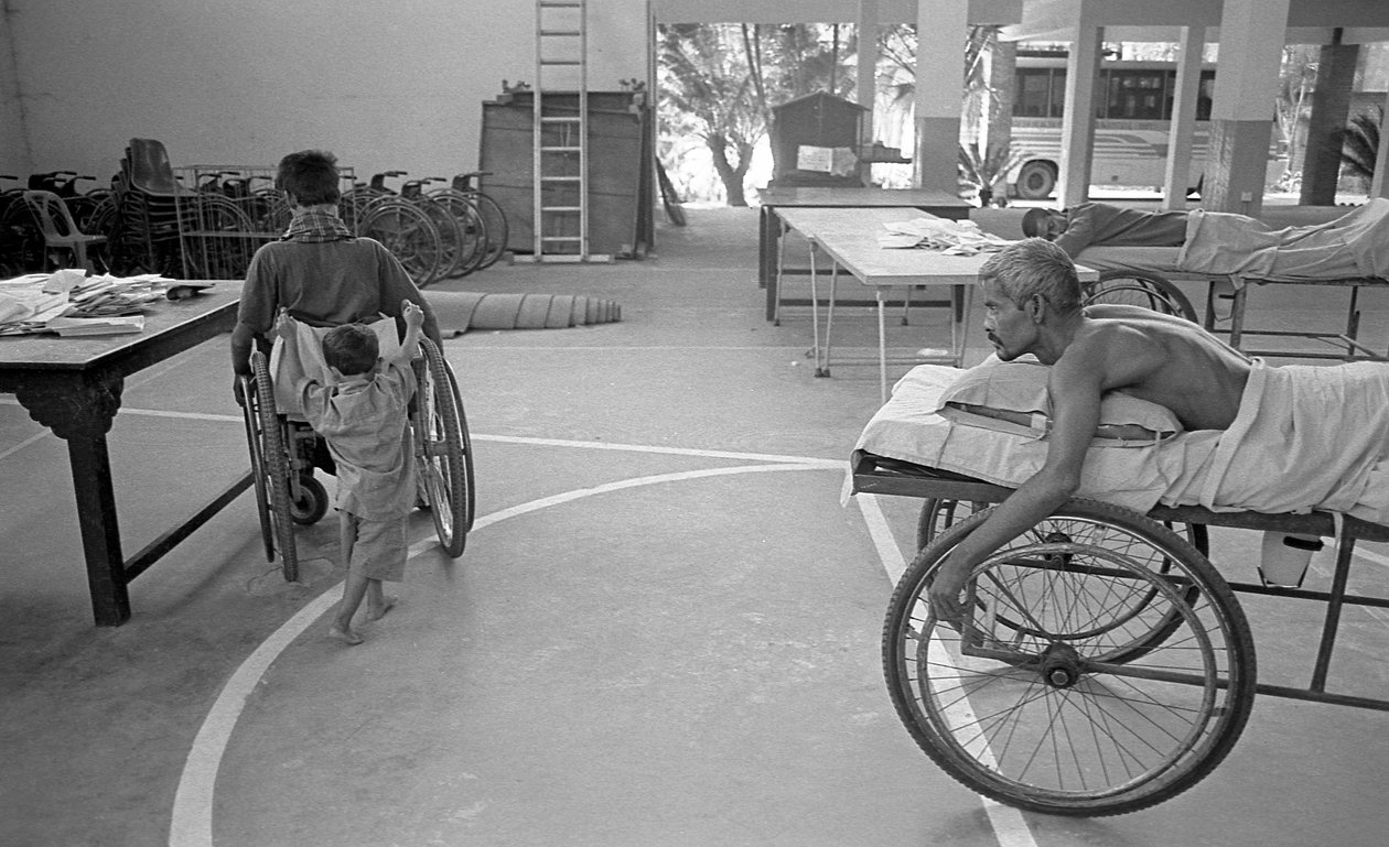 Ein körperlich behinderter Mann im Rollstuhl wird von seinem Sohn getragen; ein anderer Mann mit dem gleichen Schicksal beobachtet dies im Zentrum für die Rehabilitation der Gelähmten, CRP in Savar, Dhaka, Bangladesch. 2005. von Unbekannt Unbekannt