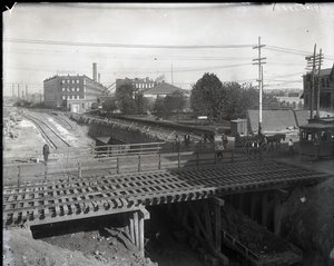 Horizontales Schwarz-Weiß-Foto einer Holzbrücke und Straßenbahnschienen über teilweise gebaute Eisenbahnschienen. Eine Straßenbahn mit der Aufschrift Chouteau 2137 beginnt rechts die Brücke zu überqueren. Im Hintergrund Fabrikgebäude mit der Aufschrift Sta