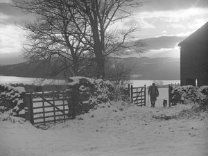 Mann und Hund gehen durch ein Tor zur Kamera im Schnee mit Thirlmere dahinter von Joseph Hardman