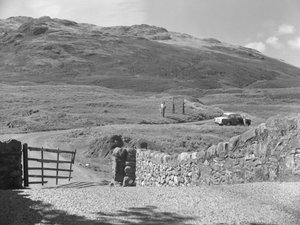 Blick auf die Straße über den Hard Knott Pass, zwei Personen stehen in der Nähe eines Straßenschilds und ein geparktes Auto auf dem Grasstreifen