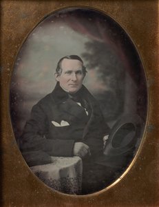 Mann mit Mantel und Handschuhen, der Hut hält, vor gemaltem Outdoor-Hintergrund sitzend von Samuel Broadbent Jr..