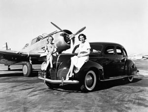 Kalifornien: 1939 Zwei Frauen sitzen auf den Kotflügeln eines 1939er Lincoln Zephyr Limousine an einem Flughafen mit einem einmotorigen Flugzeug im Hintergrund (s/w Foto) von Unbekannt