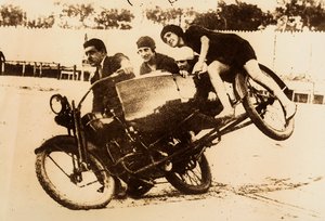 Ein Mann zeigt sich beim Fahren eines Motorrads mit Beiwagen, der drei junge Frauen trägt. Der Beiwagen hebt sich vom Boden ab, als sie eine Kurve nehmen.