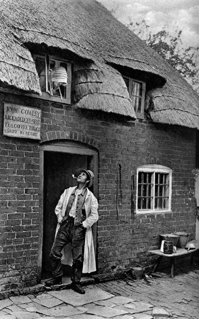 Ein Mann raucht eine Pfeife vor einem Geschäft, Worcestershire, um 1922 von A. W. Cutler