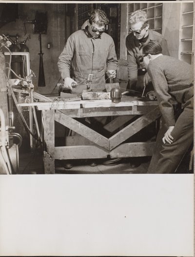 Drei Männer mit Schutzbrillen schweißen ein Metallstück auf einem Tisch von Arnold Eagle