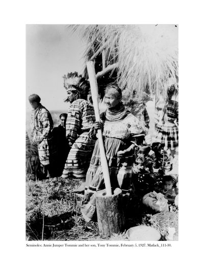 Annie Jumper Tommie benutzt einen Mörser und Stößel, um Maismehl herzustellen, und ihr Sohn, Tony Tommie, trägt einen Adlerfederkopfschmuck, 5. Februar 1927 von Claude C. Matlack