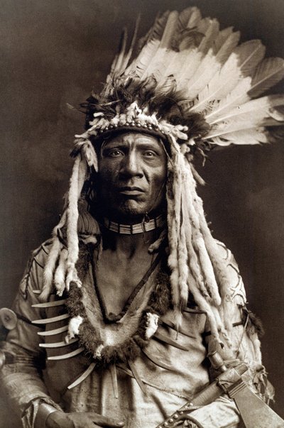 Wiesel Schwanz (Apohsuyis). Porträt des Indianerhäuptlings des Piegan-Stammes, trägt den berühmten Adlerfeder- und Wieselkopfschmuck, eine Wildledertunika, zwei Halsketten, eine aus Knochen und die andere aus Grizzlyklauen, und eine Tomahawk-Pfeife von Edward Sheriff Curtis