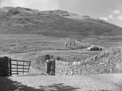 Blick auf die Straße über den Hard Knott Pass, zwei Personen stehen in der Nähe eines Straßenschilds und ein geparktes Auto auf dem Grasstreifen von Joseph Hardman