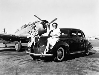 Kalifornien: 1939 Zwei Frauen sitzen auf den Kotflügeln eines 1939er Lincoln Zephyr Limousine an einem Flughafen mit einem einmotorigen Flugzeug im Hintergrund (s/w Foto) von Unbekannt