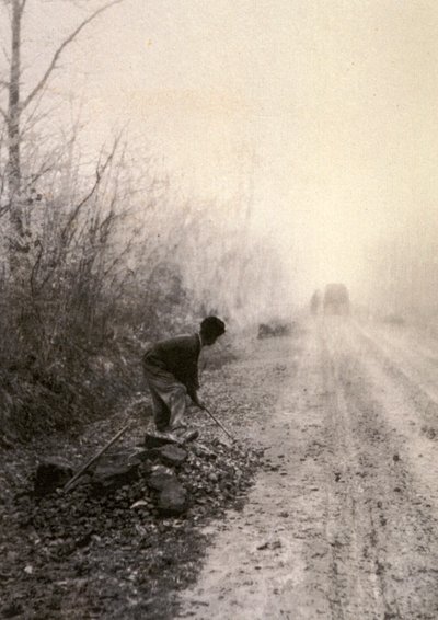 Eine Landstraße in Morgennebel gehüllt. Im Vordergrund spaltet ein Mann Steine von Unbekannter Künstler