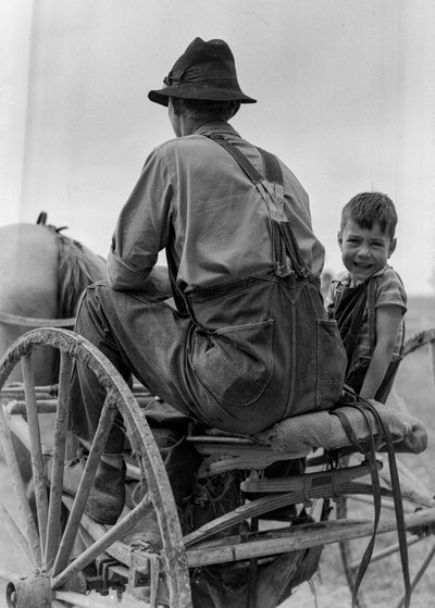 Bauer und lächelnder Junge fahren auf dem Sitz eines pferdegezogenen Wagens in West Virginia von Volkmar Wentzel