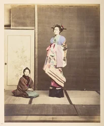 [?Geisha,] Ganzkörperporträt einer jungen Frau in erhöhten Schuhen, ihr junger Begleiter kniet neben ihr