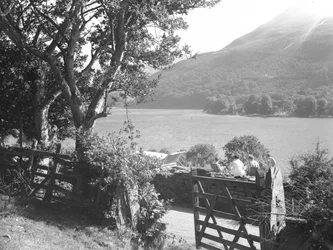 Vier Personen sitzen auf einer Trockenmauer und machen ein Picknick mit Blick auf den See, Auto mit offenem Kofferraum geparkt