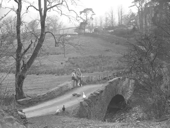 Zwei Mädchen sitzen auf einer Steinbrücke über den Fluss Cocker am Fuße des Scale Hill und beobachten Gänse auf dem Weg bei Loweswater